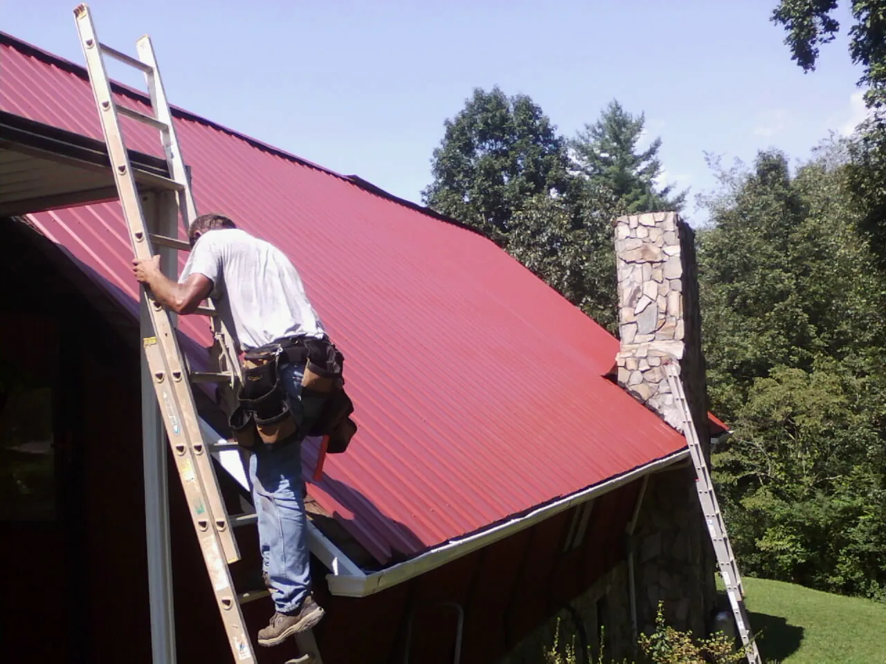 Roofer on a ladder beside a red metal roof and stone chimney — Soffit & Fascia Repair by Roofing Brothers in Kenton